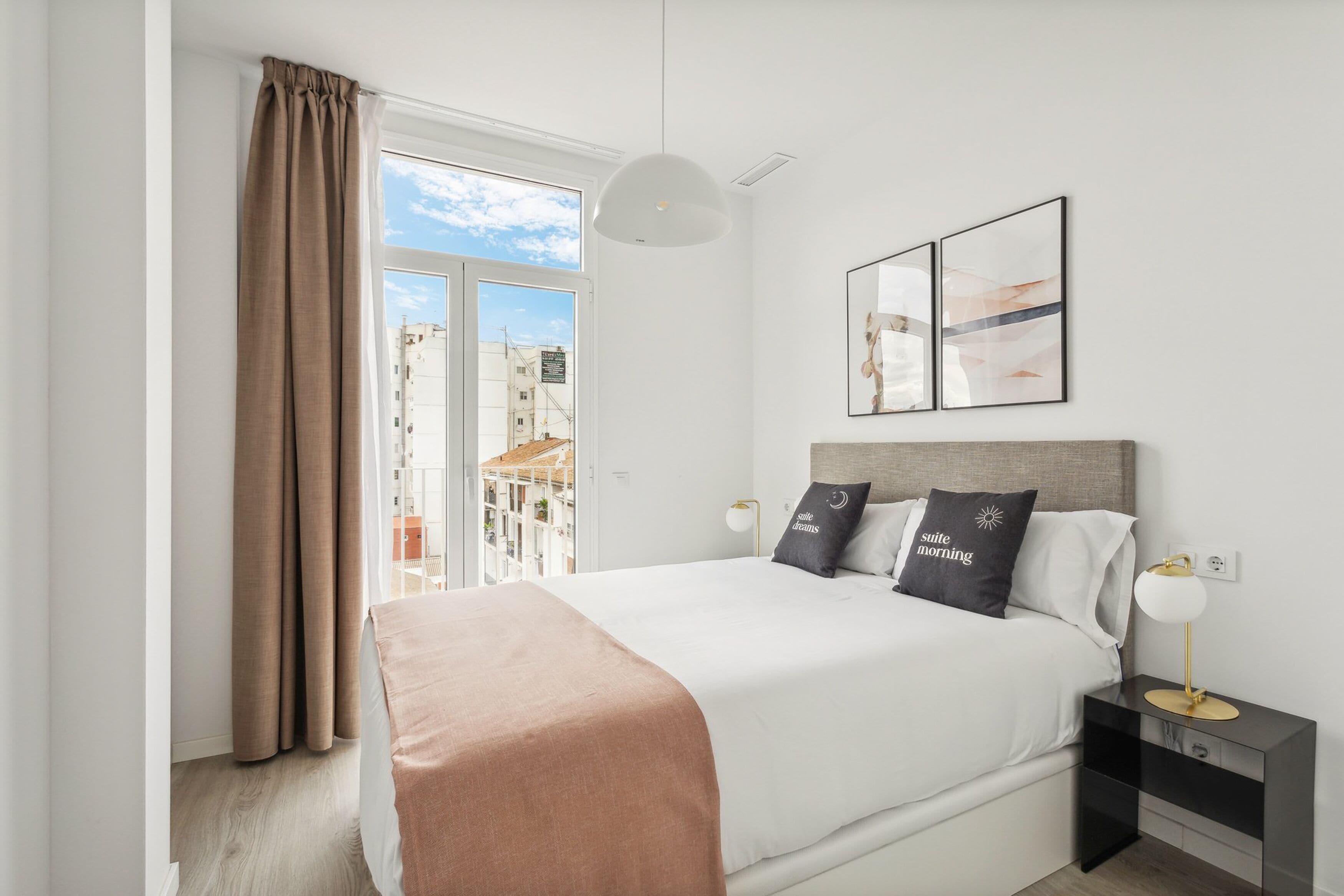A modern hotel room with a large bed, a bedside table with a lamp, and a view of a cityscape through a large window.