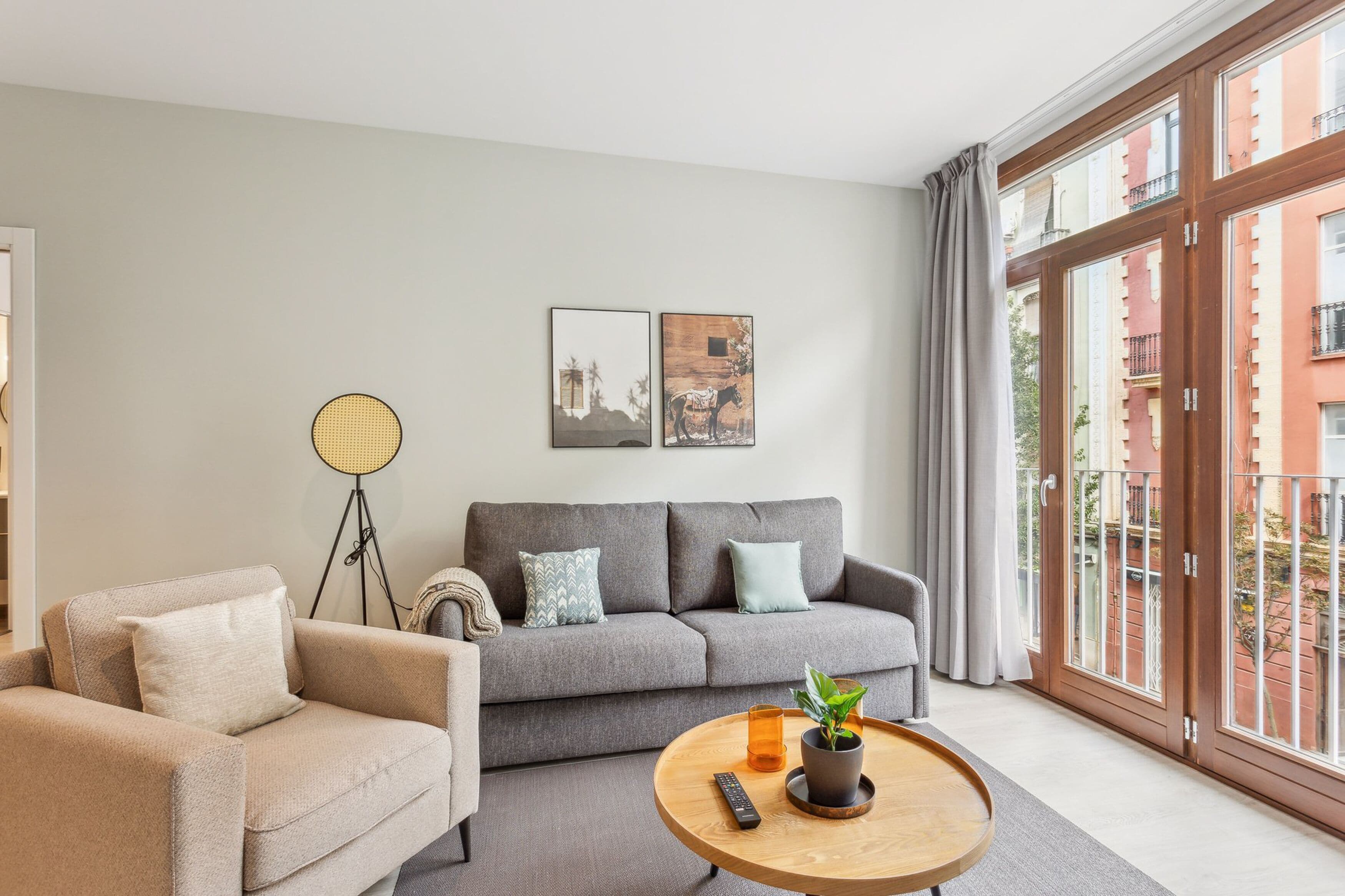 A modern living room with a grey sofa, beige armchair, and a round wooden coffee table. There are two framed pictures on the wall, a floor lamp, and a large window with a view of buildings.