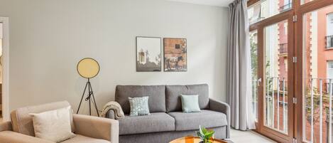 A modern living room with a grey sofa, beige armchair, and a round wooden coffee table. There are two framed pictures on the wall, a floor lamp, and a large window with a view of buildings.