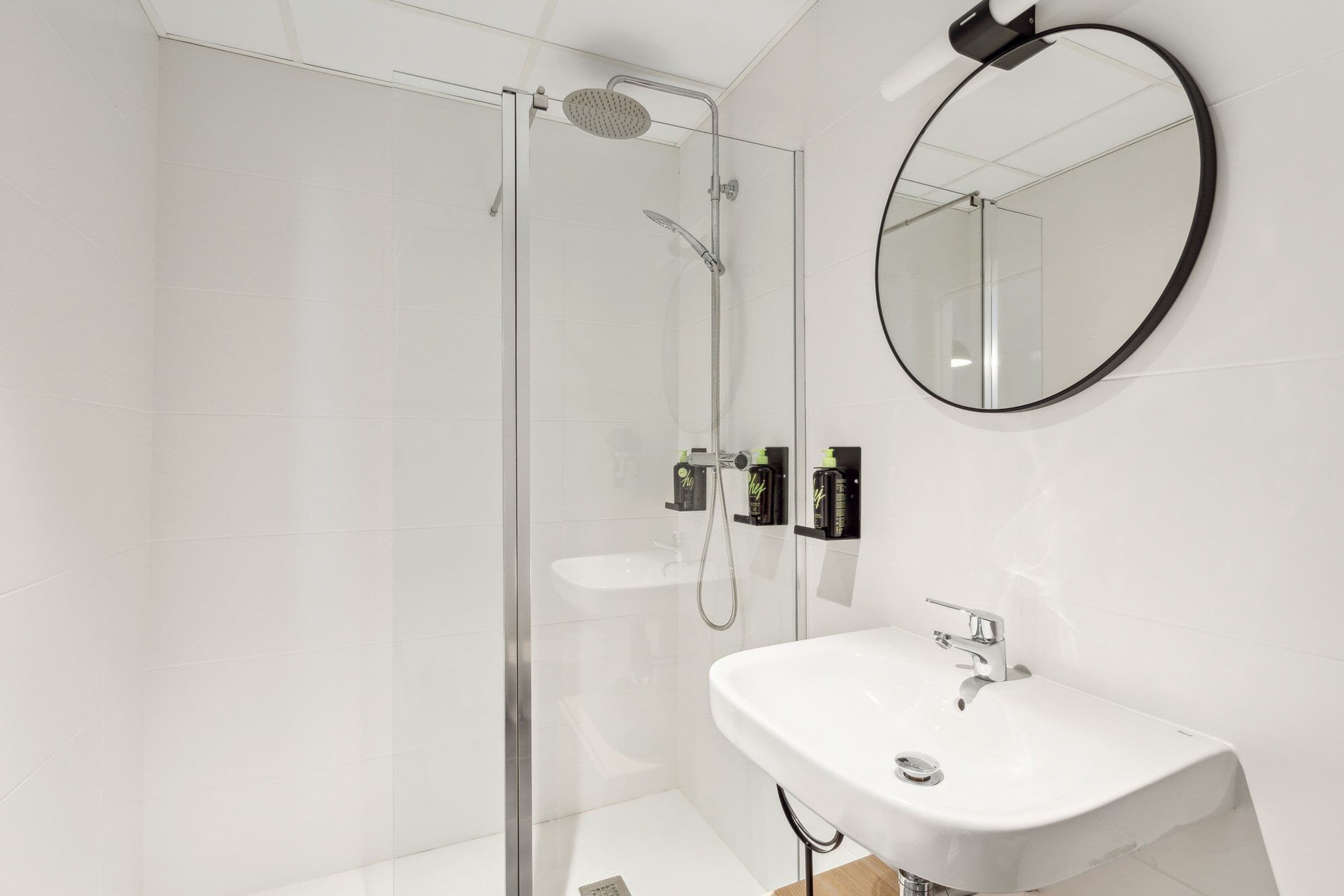 Un baño moderno con ducha envuelta en vidrio, lavamanos blanco y espejo redondo en la pared.