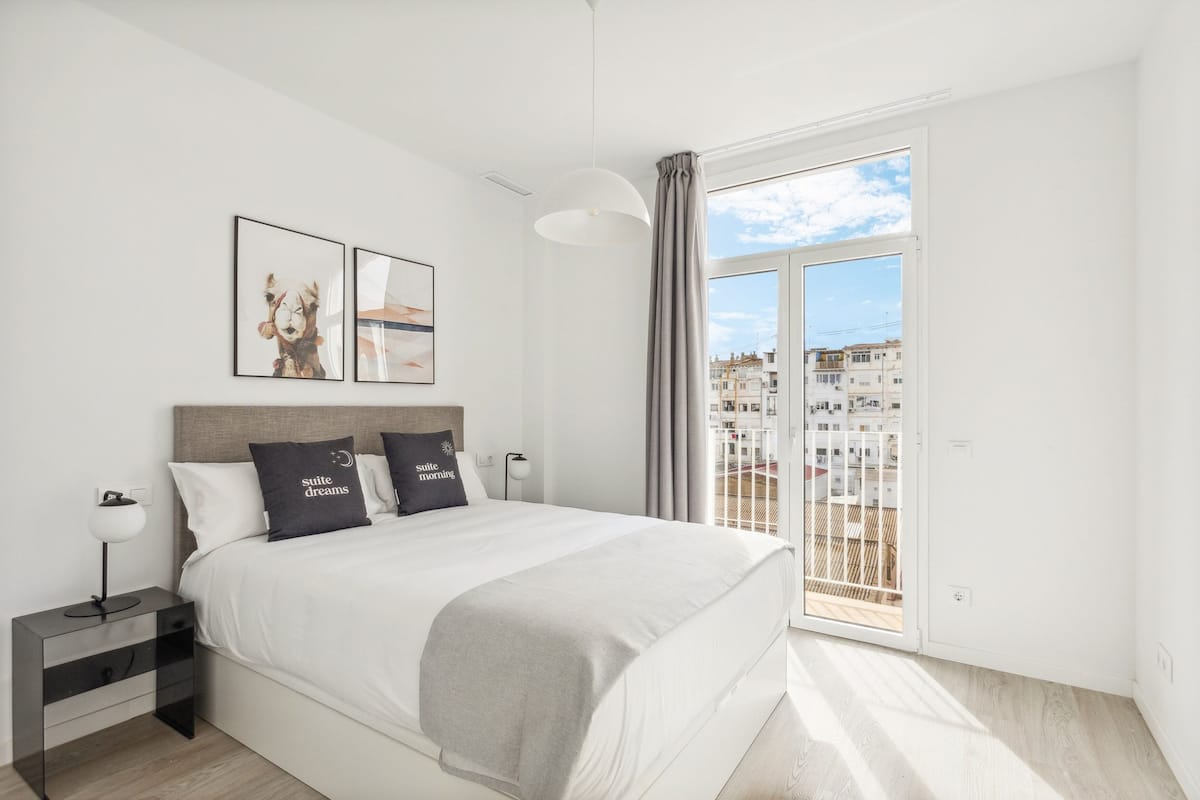 A modern bedroom with a large bed, a nightstand, and a view of a cityscape through a sliding glass door.
