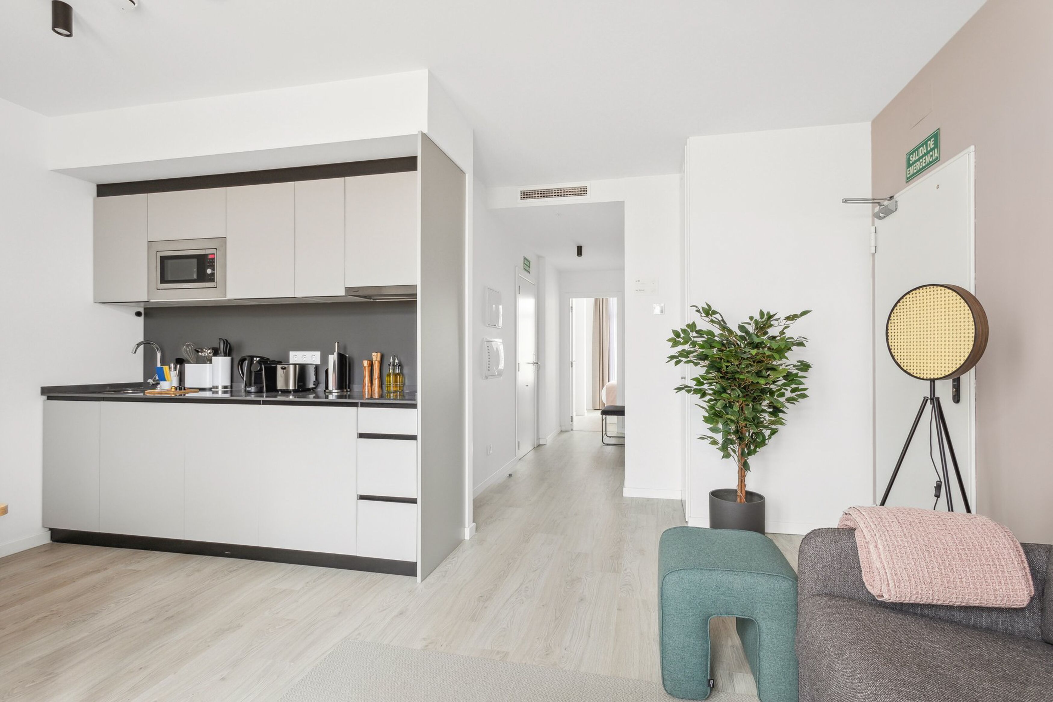 Une cuisine moderne avec des meubles blancs, un four à micro-ondes et une machine à café. On y trouve une plante en pot et une table ronde avec une chaise en osier. Un canapé gris avec un plaid rose est visible.