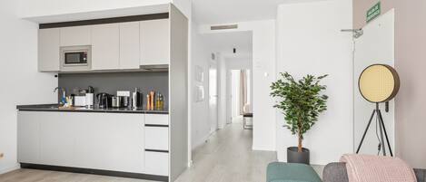 Une cuisine moderne avec des meubles blancs, un four à micro-ondes et une machine à café. On y trouve une plante en pot et une table ronde avec une chaise en osier. Un canapé gris avec un plaid rose est visible.