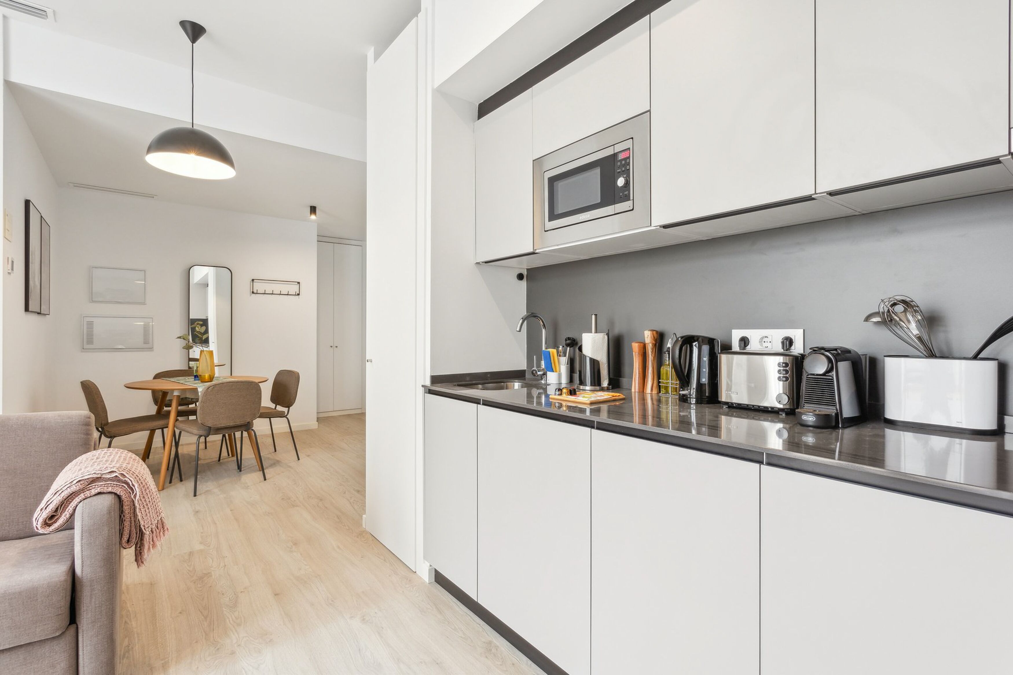 A modern kitchen with white cabinets, a microwave, a toaster, and a coffee maker. There is a dining area with a table and chairs.