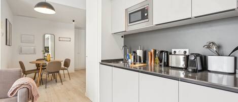 A modern kitchen with white cabinets, a microwave, a toaster, and a coffee maker. There is a dining area with a table and chairs.