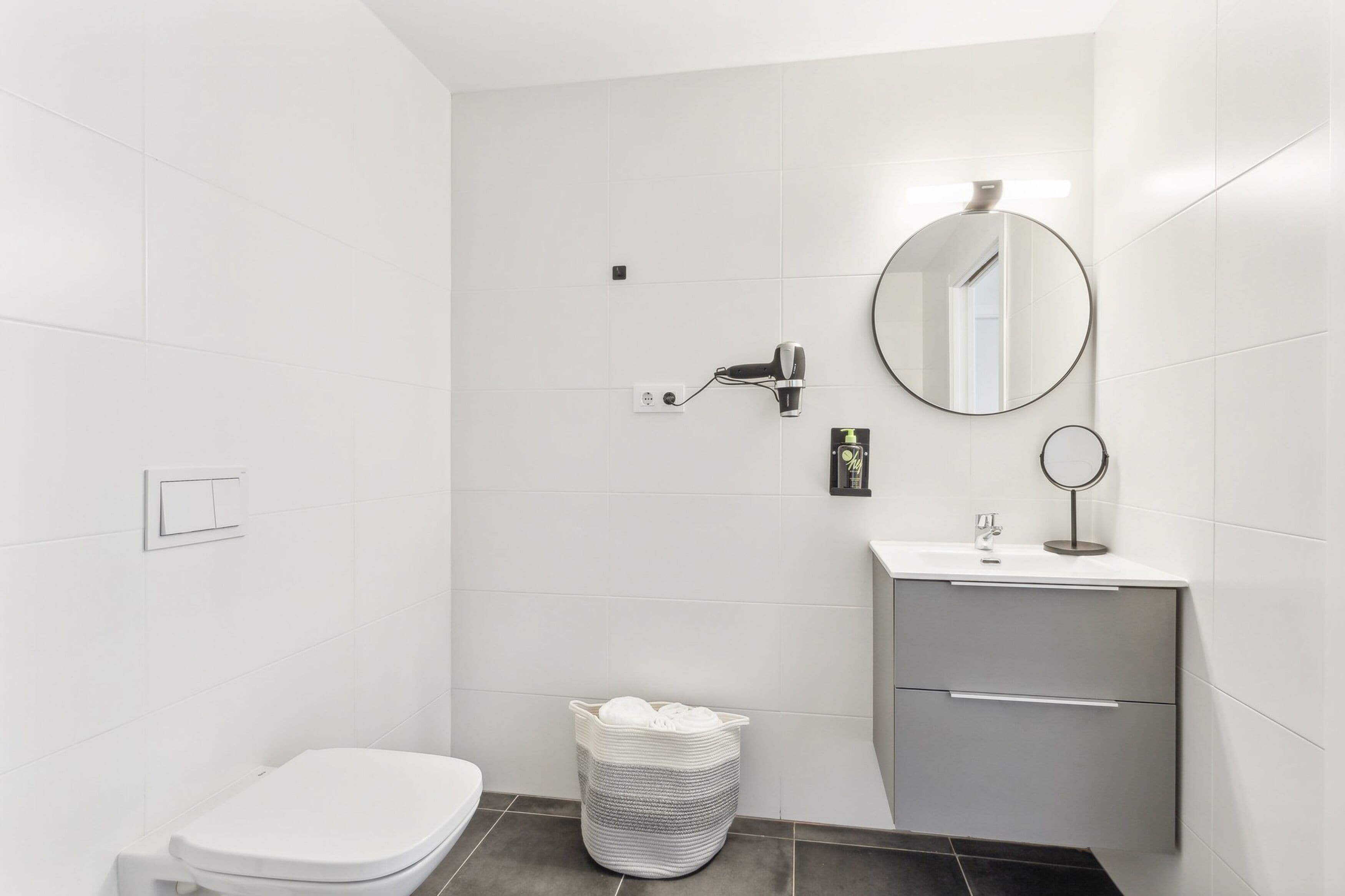 A modern bathroom with white tiles, a toilet, a sink with a mirror, and a towel basket.