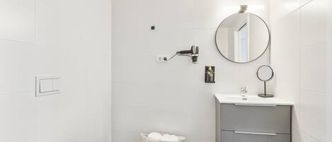 A modern bathroom with white tiles, a toilet, a sink with a mirror, and a towel basket.