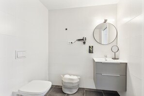 A modern bathroom with white tiles, a toilet, a sink with a mirror, and a towel basket. - Limehome Valencia Calle de Cadiz (Valencia)