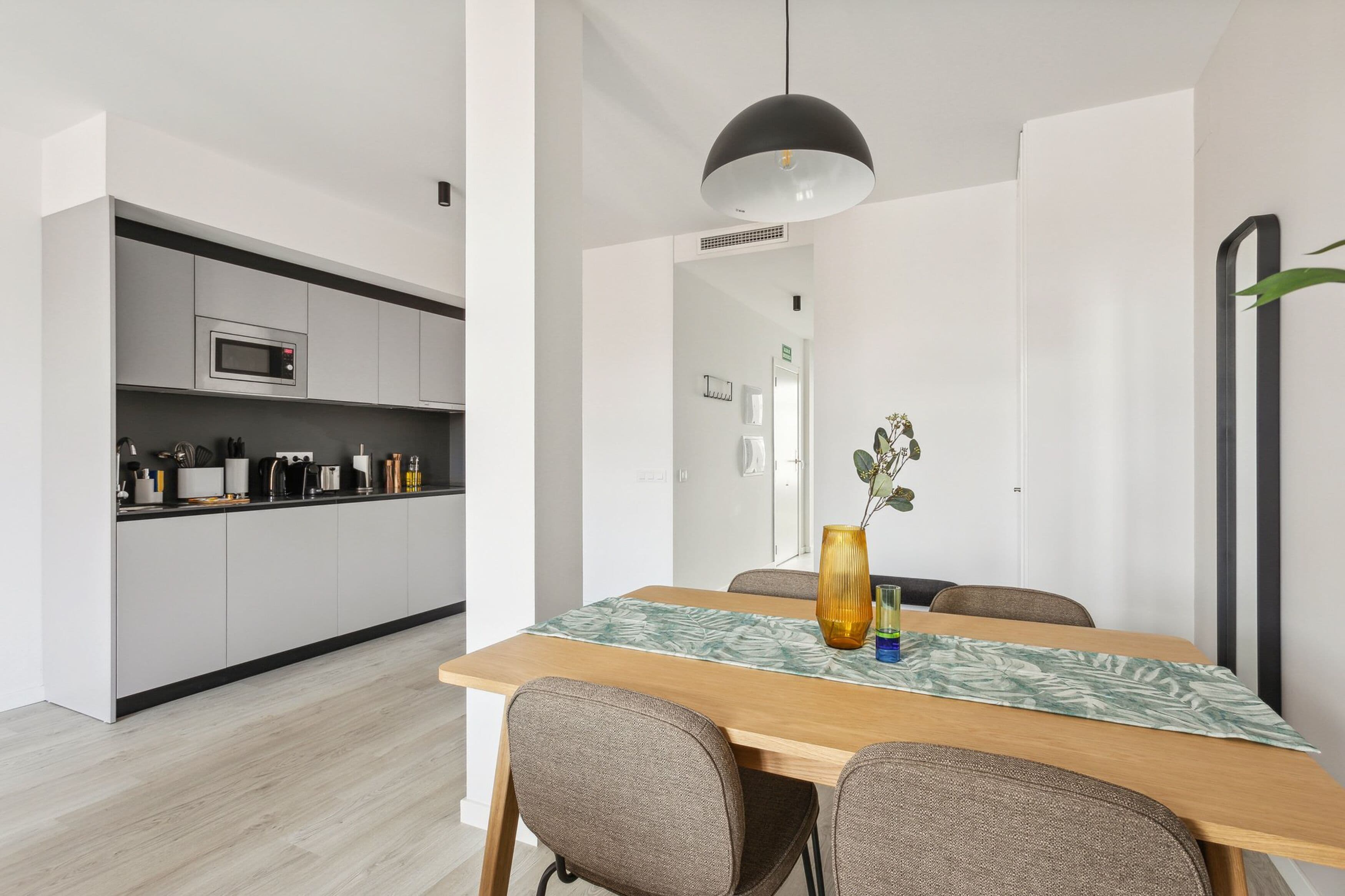 A modern kitchen with white cabinets and a microwave, a dining area with a wooden table and chairs, and a pendant light above.