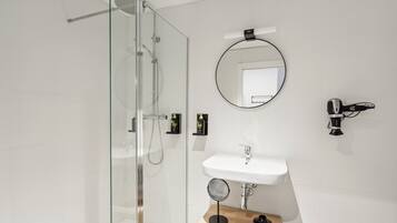 A modern bathroom with a glass shower enclosure, a white sink, a round mirror, and a hairdryer mounted on the wall.
