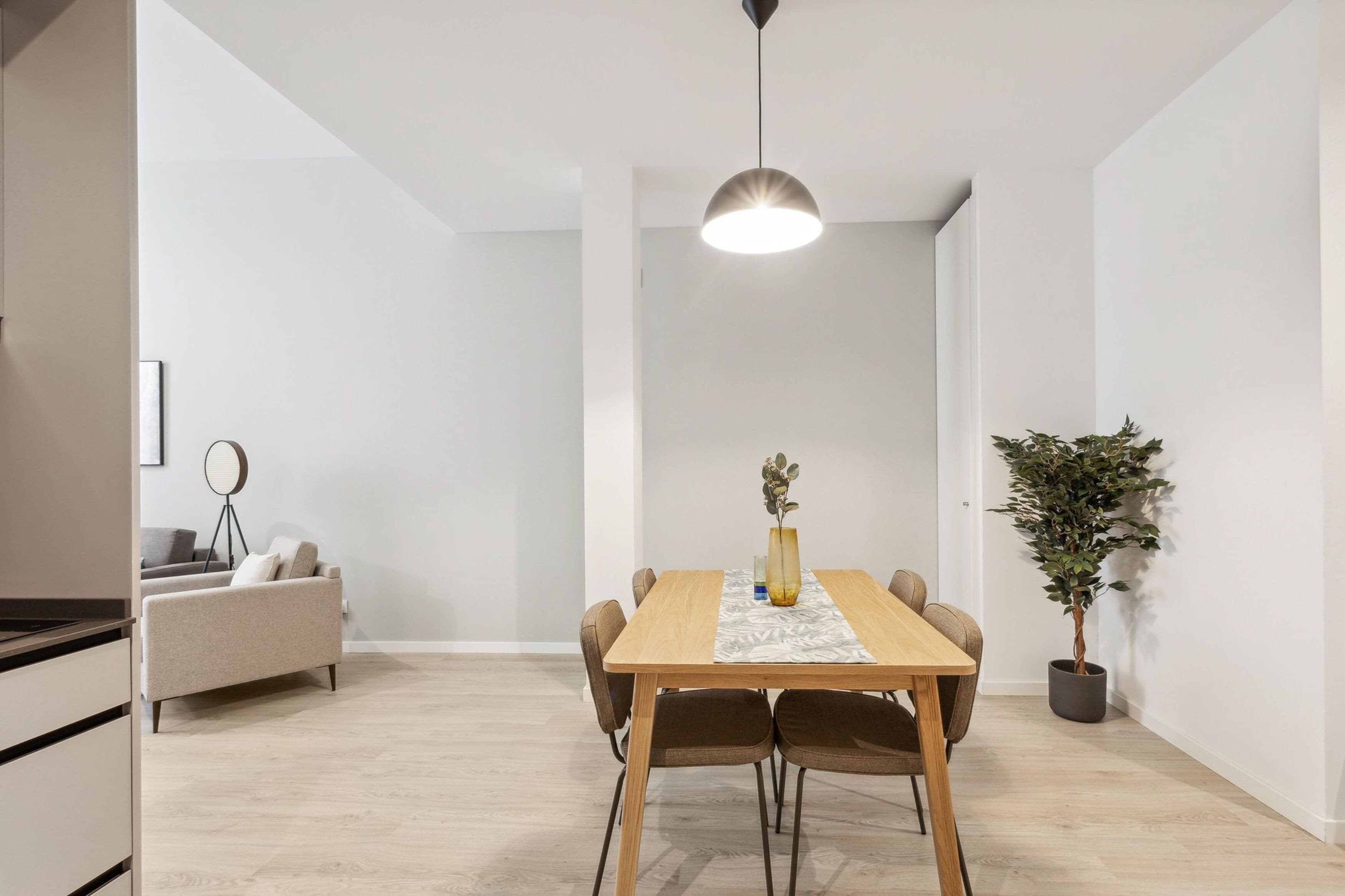 A modern dining area with a wooden table, six chairs, a vase with flowers, a hanging light fixture, a sofa, and a potted plant.