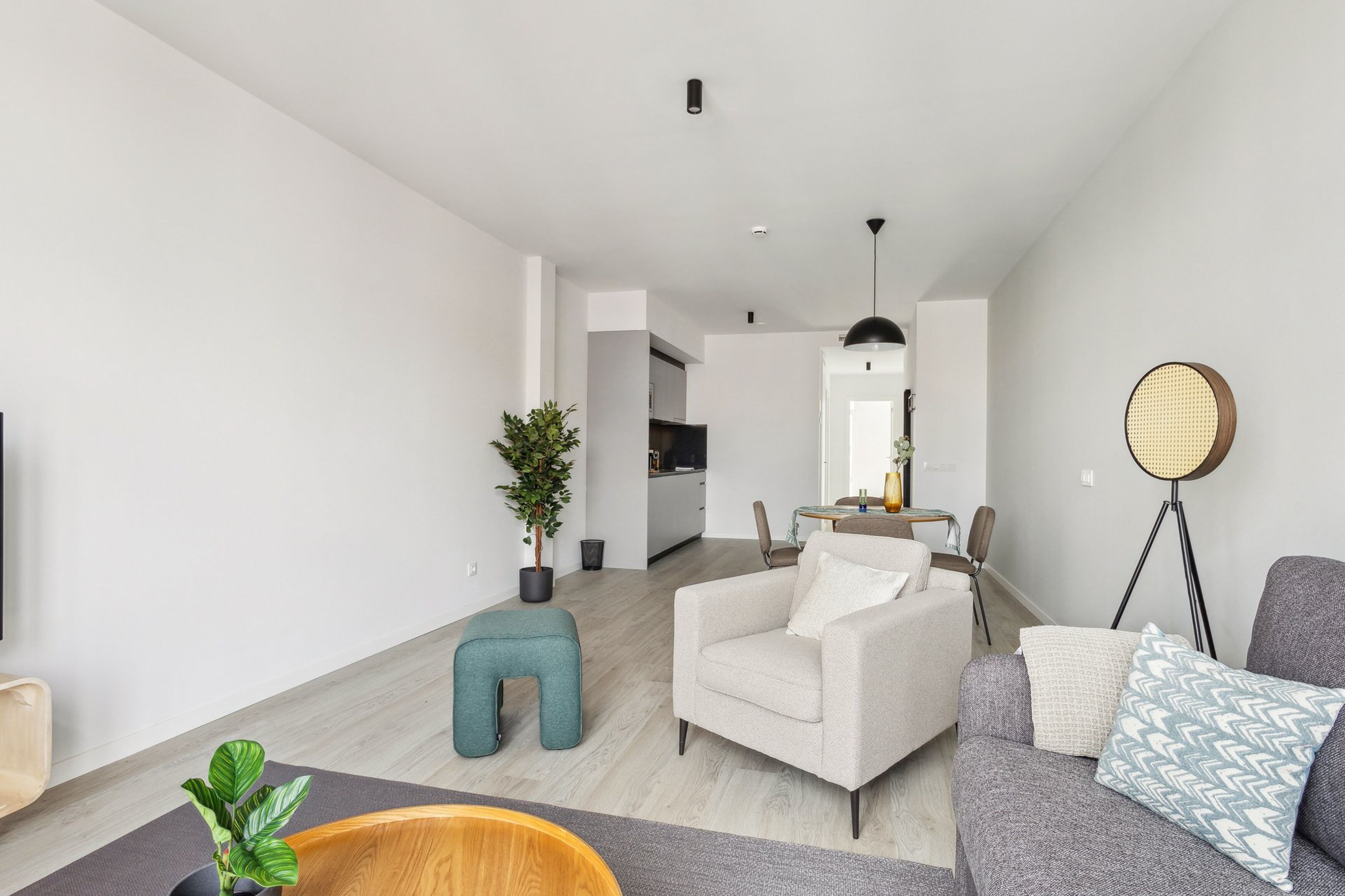 A modern living room with a sofa, armchair, and ottoman, a dining area with a table and chairs, and a kitchen in the background.
