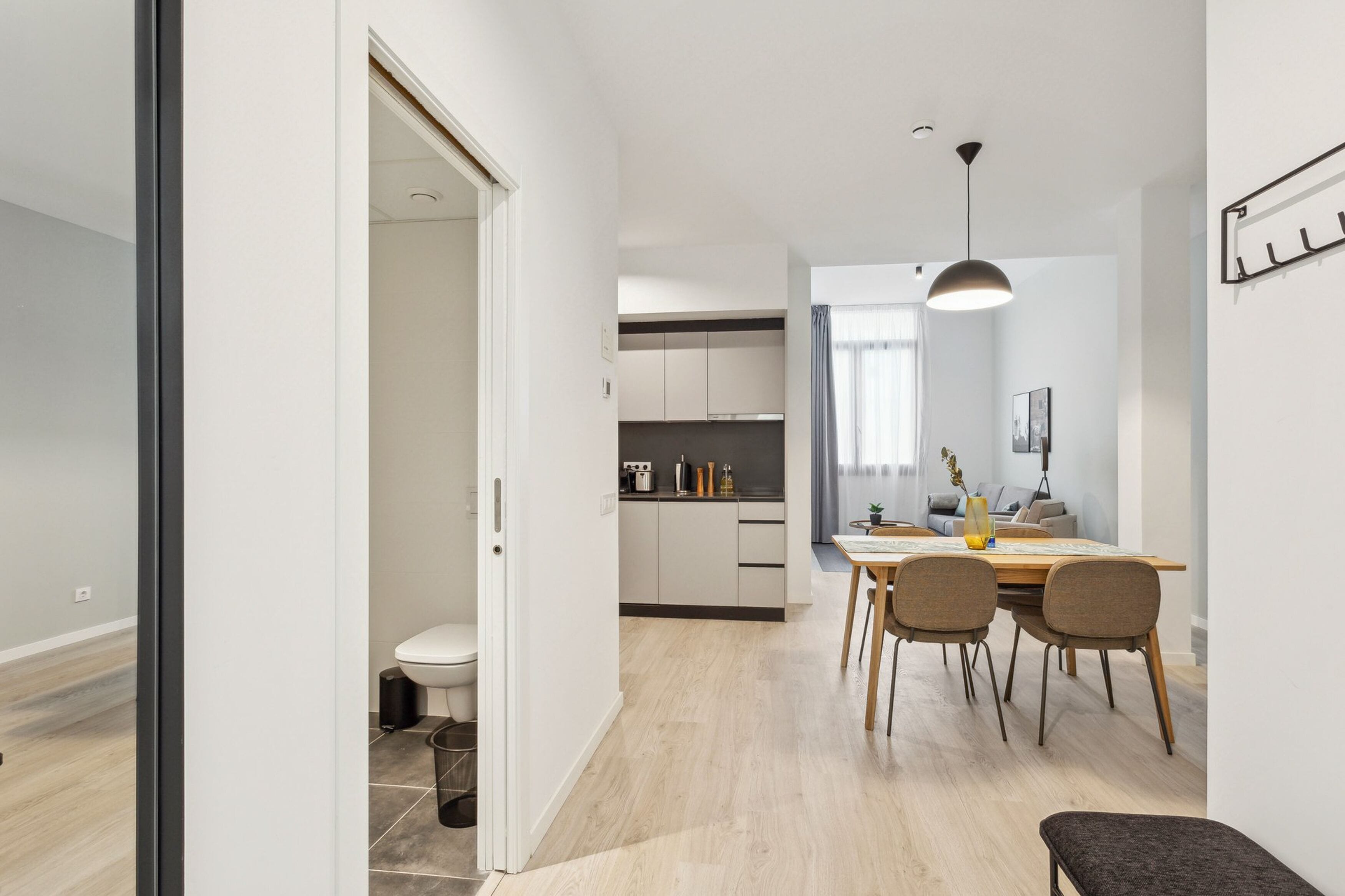 Una cocina moderna con armarios blancos, un comedor con mesa y sillas de madera, y un baño visible con inodoro y lavamanos.