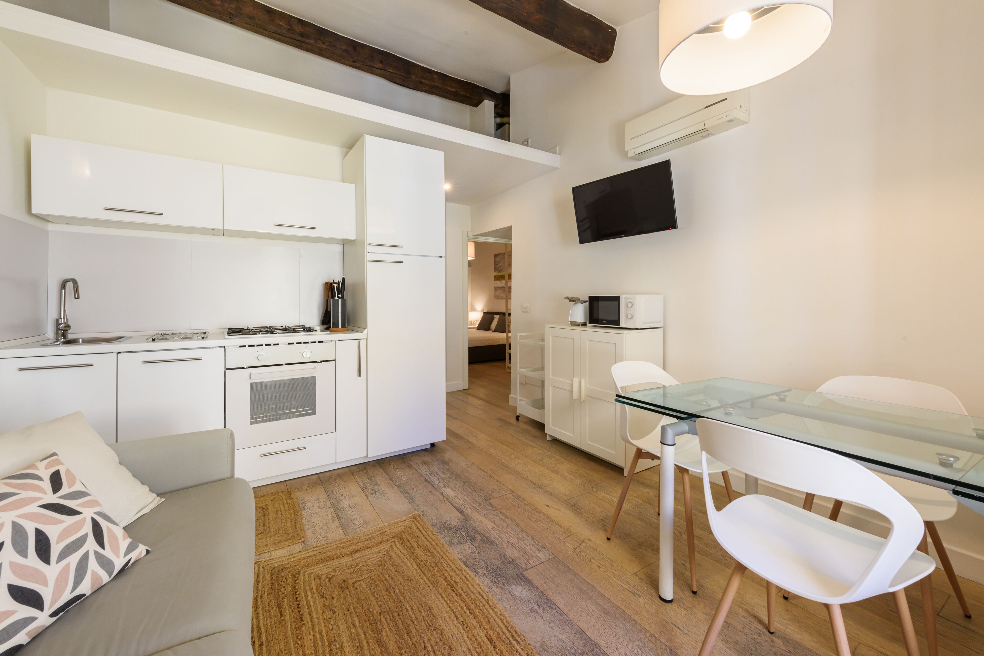 Una cocina moderna con armarios blancos, un comedor con mesa de vidrio y sillas blancas, y un televisor de pantalla plana montado en la pared.