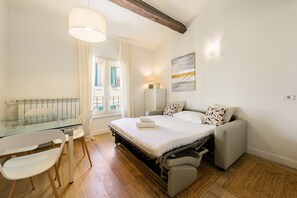 A bedroom with a glass dining table, a sofa, a bed with a mattress and pillows, a bedside table with a lamp, and a window with curtains. - Novella Contemporary Apartment by Mmega (Florence)