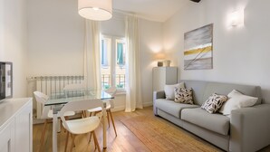 A modern living room with a glass dining table, white chairs, a grey sofa with patterned cushions, a painting on the wall, and a ceiling light fixture. - Novella Contemporary Apartment by Mmega (Florence)