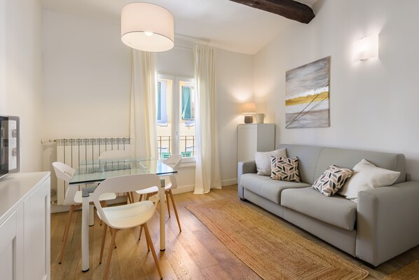 A modern living room with a glass dining table, white chairs, a grey sofa with patterned cushions, a painting on the wall, and a ceiling light fixture. - Novella Contemporary Apartment by Mmega (Florence)