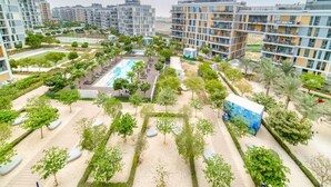 A modern residential complex with multiple high-rise buildings, a central swimming pool, landscaped gardens, and a variety of trees.