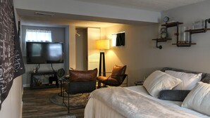 A hotel room with a bed, a chair, a TV, a lamp, and shelves with items.