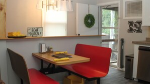 A compact kitchen with a dining area, a sign reading "SEAT YOURSELF", a red chair, a small table, and a yellow stool.