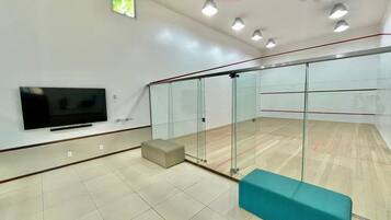 A squash court with glass walls, a television, and a bench.