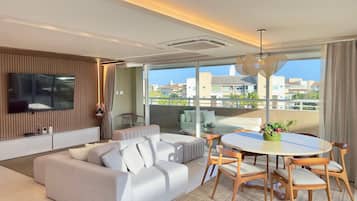 Ein modernes Wohnzimmer mit einer Couch, einem Esstisch mit Stühlen, einem an der Wand montierten Fernseher und einem Balkon mit Blick auf Gebäude.