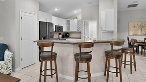 Una cocina moderna con armarios blancos, una isla central con taburetes y electrodomésticos como refrigerador y microondas.
