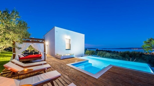 A modern outdoor pool area with a wooden deck, lounge chairs, and a small building with a window. - Dalmatian Oasis Luxury Villa (Kaštel Lukšić)