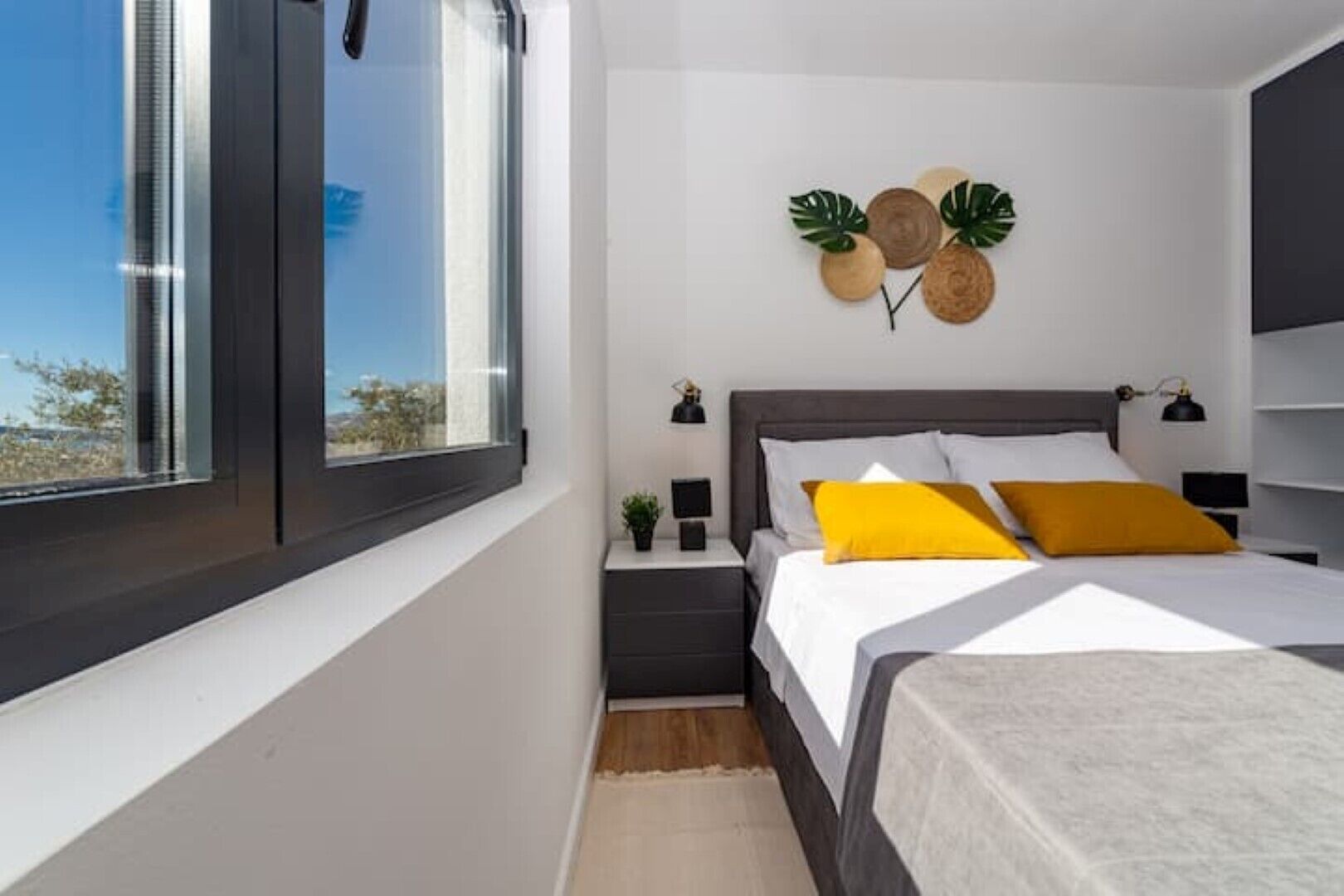 Un dormitorio moderno con un ventanal grande, una cama con ropa de cama blanca y amarilla, una mesita de noche con una lámpara, y detalles decorativos en la pared que incluyen una planta y un objeto de madera.