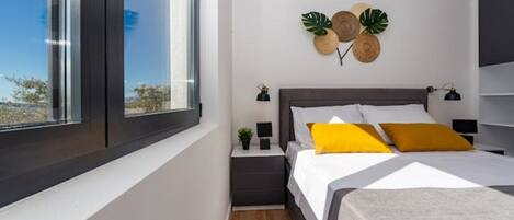 Un dormitorio moderno con un ventanal grande, una cama con ropa de cama blanca y amarilla, una mesita de noche con una lámpara, y detalles decorativos en la pared que incluyen una planta y un objeto de madera.