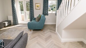 A spacious living room with a herringbone floor, a teal armchair, a grey sofa, and a staircase leading to an upper floor.