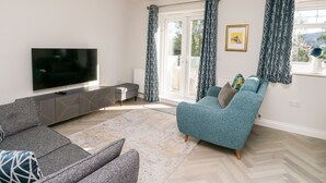 A modern living room with a flat-screen TV, grey sofa, teal armchair, patterned curtains, and a decorative ceiling light.