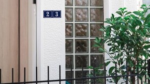 Entrada de un edificio con puerta de madera, un cartel con el número 23, un ventanal con vidrio esmerilado y una planta en maceta.
