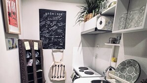 Una cocina compacta con un lavamanos, estufa y estanterías. En la pared hay una pizarra con escritura, y hay una planta en la estantería.