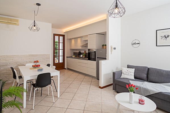 A modern kitchen with a dining area, a grey sofa, and a small table with a vase of flowers.
