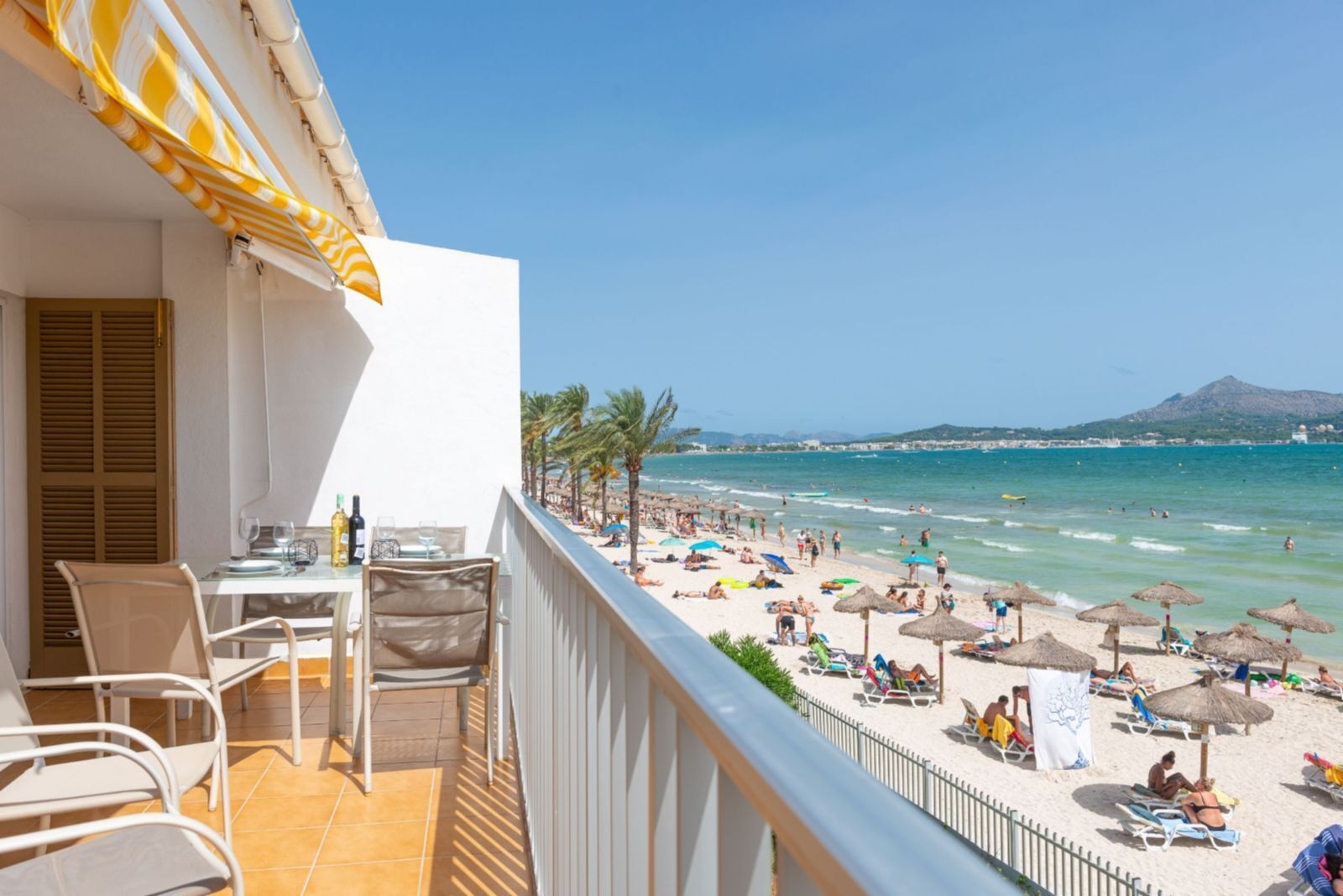 A balcony with a table set for dining, overlooking a beach with umbrellas and people.