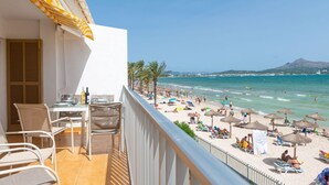 Balcón con mesa lista para comer, con vistas a una playa con sombrillas y gente.