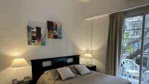 Une chambre à coucher avec un lit, deux tables de chevet avec des lampes, une fenêtre donnant sur l’extérieur et deux tableaux abstraits accrochés au mur.