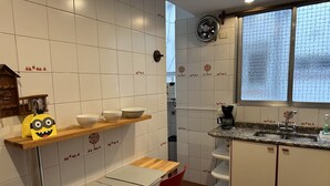 Une cuisine avec des murs recouverts de carreaux blancs, une étagère en bois avec des bols, une chaise rouge, une machine à café et une fenêtre donnant sur des arbres.