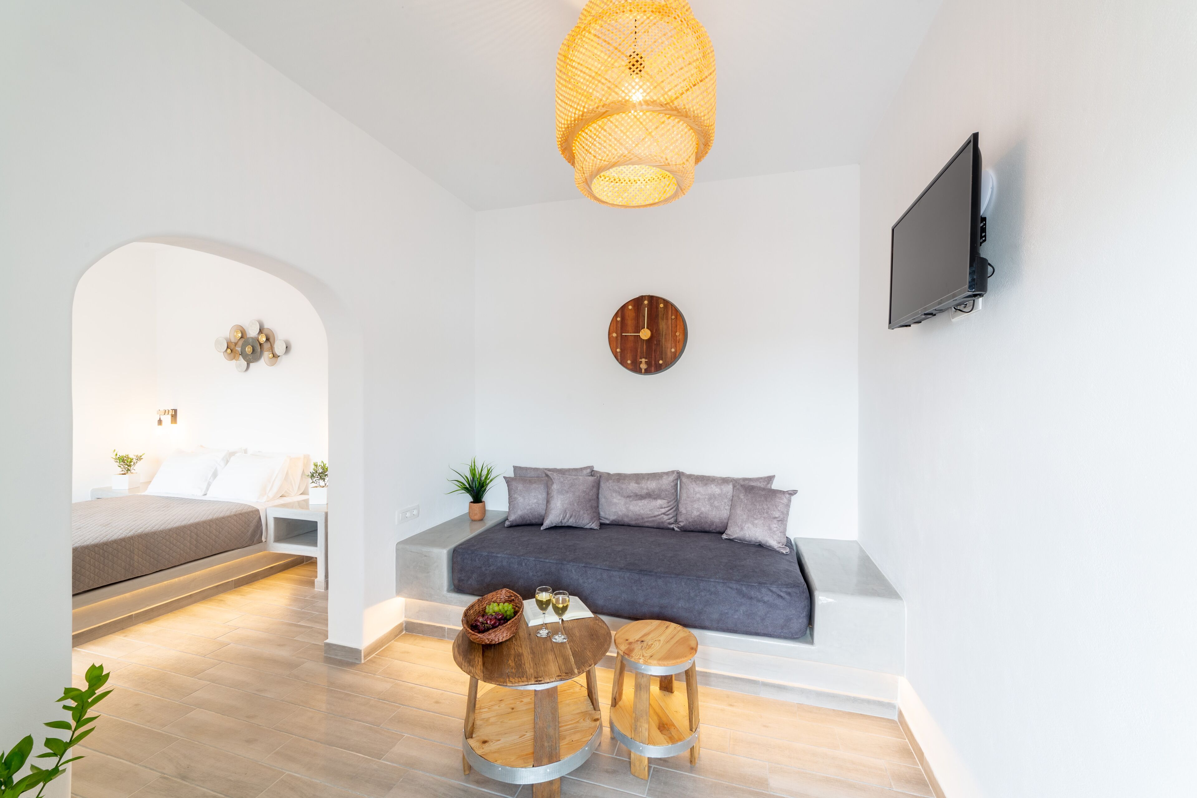 Un soggiorno moderno con un divano, due sgabelli in legno, un tavolino con un cesto di frutta, una TV a parete e un originale lampadario intrecciato.