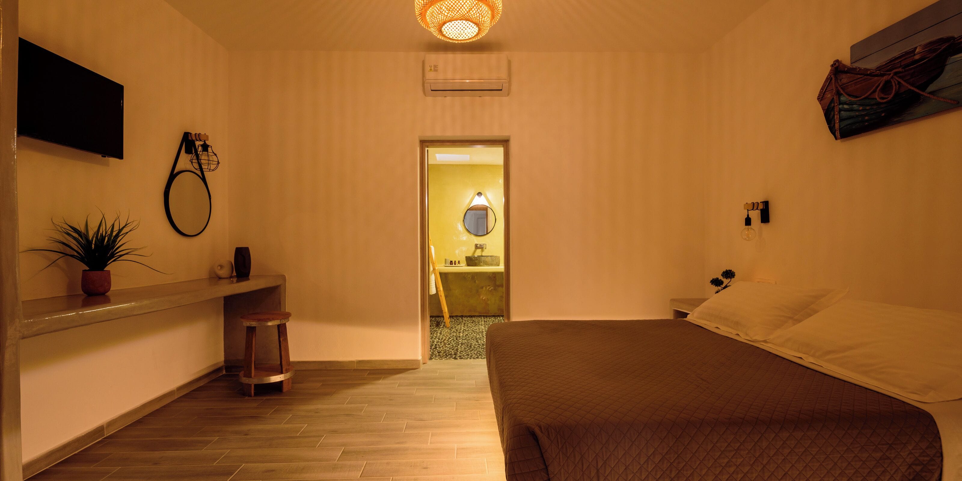 Habitación de hotel con cama, un escritorio pequeño con silla, una planta en maceta, un televisor montado en la pared y una puerta que da a un baño.
