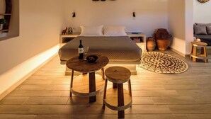 A spacious bedroom with a large bed, a bedside table with a bottle and glass, a small round table with a bowl, and a woven light fixture above. - Athiri Luxury Suites Perivolos (Santorini)