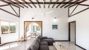 Un vaste espace de vie comprenant un grand canapé d’angle gris, une table basse en bois et un ventilateur de plafond. On aperçoit un coin repas à travers une arche, et un tableau orne le mur.