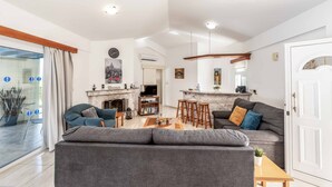 Una sala de estar moderna con sofá, mesa de centro y taburetes de bar. Se ve una chimenea, un televisor y una zona de cocina.