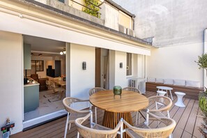 Um deck de madeira com uma mesa redonda e cadeiras de vime, com vista para um interior moderno com sala de jantar e cozinha.