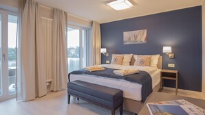 A hotel room with a bed, bench, bedside tables, and a view of the outdoors. - Dünenresidenz Prora (Binz)