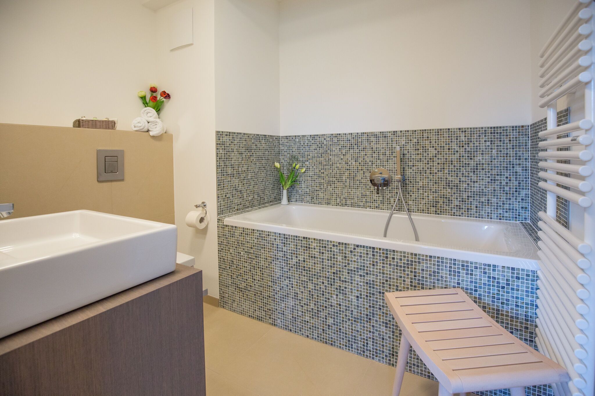 Une salle de bain avec une grande baignoire rectangulaire, un lavabo blanc et un petit tabouret en bois.