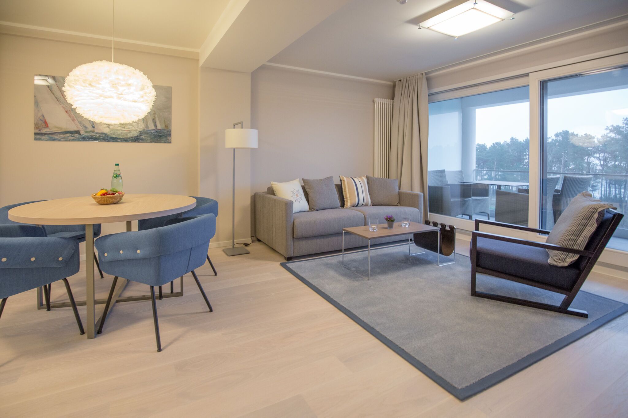Un salon moderne avec une table à manger ronde, des chaises bleues, un canapé gris, une table basse et une grande fenêtre donnant sur un beau paysage.