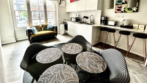 A modern kitchen with white cabinets, a microwave, and a coffee machine. There is a dining area with black chairs and patterned table mats. - soho london suite (London)