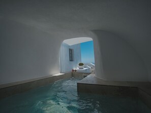 A narrow, water-filled passageway with a clear view of a white room with a window and a plant. - Anemos Family Suites (Santorini)